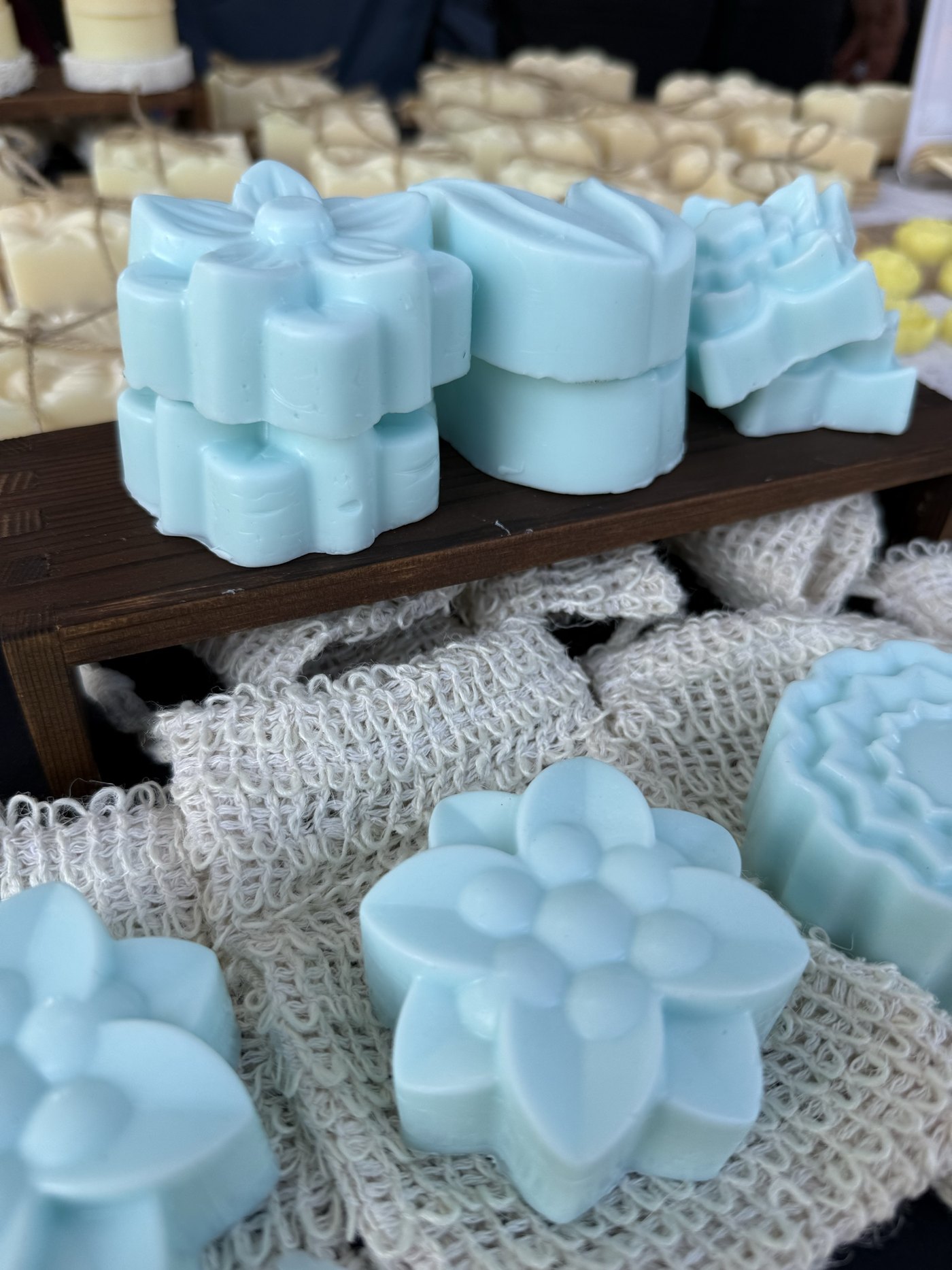 Pale blue flower-shaped handmade soaps stacked on a wooden riser beside woven soap saver cloths